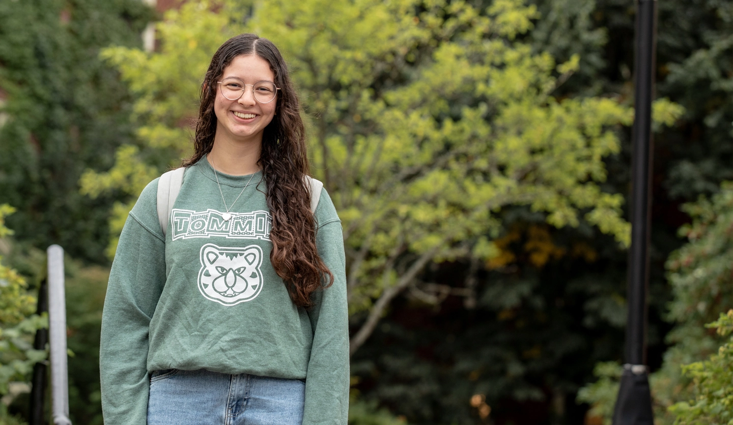 A female student wearing a Tommies sweater stands smiling outside on campus