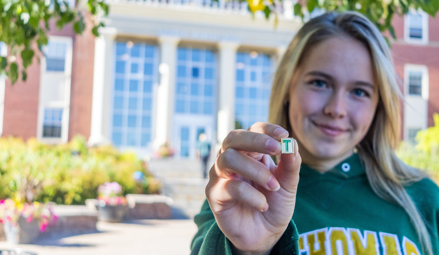 A female student wearing a green St. Thomas hoodie holds out a gold T-pin