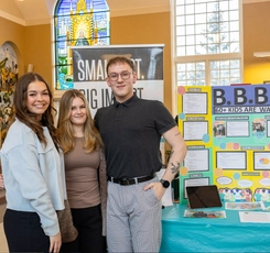 Two female students and one male student standing similing in front of a banner for the Big Brothers Big Sisters of Fredericton and Oromocto