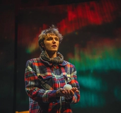 A male actor in a plaid coat holds a fake snowball in front of the northern lights in the Black Box Theatre