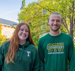 A female student with red hair and a green hoodie stands beside a male student with blonde hair wearing a STU Tommies Soccer shirt