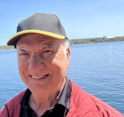 Doug Dolan stands in a red coat and blue baseball cap on the shore of the Miramichi River