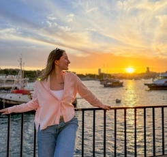 Kamylle leaning against a waterfront railing in Malta, looking toward a golden sunset over the harbour, with boats and buildings in the background.