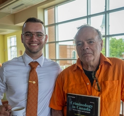 Simon Wassef and his Dad, Chris McCormick, stand together in the Study Hall. Simon holds a cup of coffee and Chris is holding a Criminology in Canada textbook.