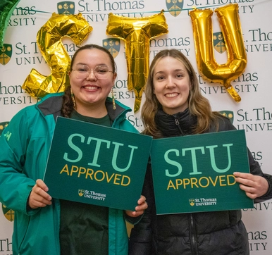 Two female students stand in front of a St. Thomas University backdrop holding signs that read 