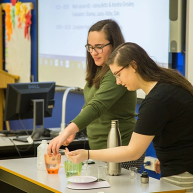Two women standing and conducting experiments in a classroom with beakers, weighing machine and a water bottle laid out in table