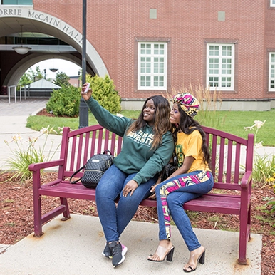Two students sitting on a bench and taking a selfie outside of Margaret Norrie McCain Hall
