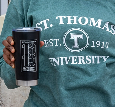 A student wearing a green STU sweater while holding up a St. Thomas University tumbler