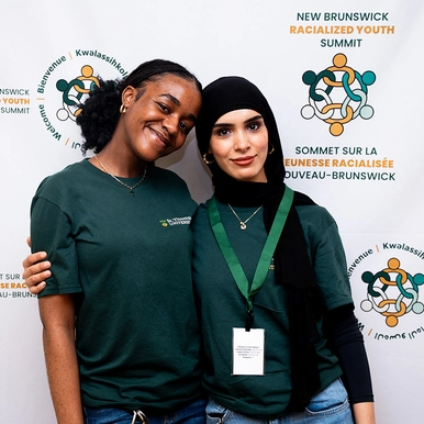Two female ������������ students standing in front of NB Racialized Youth Summit banner