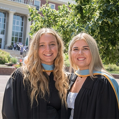 Two female Bachelor of Education students wearing their blue and gold laced gowns on campus next to GMH