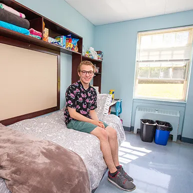 A student sitting on bed in dorm room.