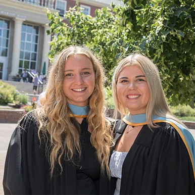 Two female Bachelor of Education students wearing their blue and gold laced gowns on campus next to GMH