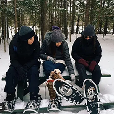 Three students wearing snowshoes sitting in the forest.