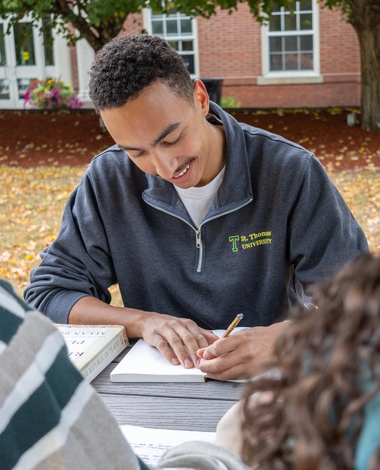 A male student wearing a STU sweater sits at a table in the lower courtyard doing school work