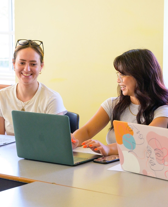Two female students sit at a table in class with their laptops out