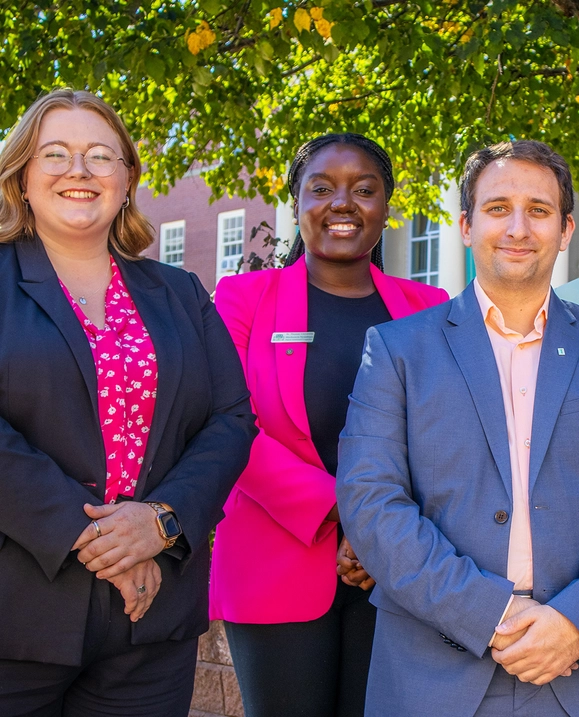 Two women and a man stand side by side outside on campus