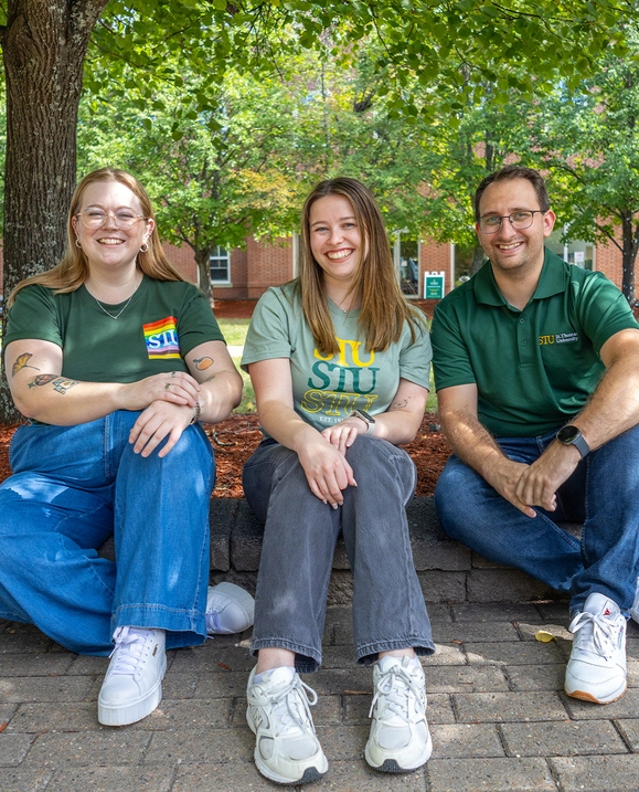 Three Admissions Counsellors—two female and one male—sit together under a tree on campus