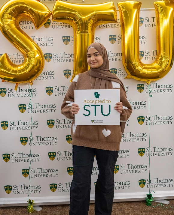 A female student stands in front of a St. Thomas University backdrop holding a sign that reads 