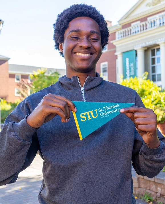 A male student wearing a gray sweatshirt holds a �ǿմ�ý pennant in front of George Martin Hall on campus