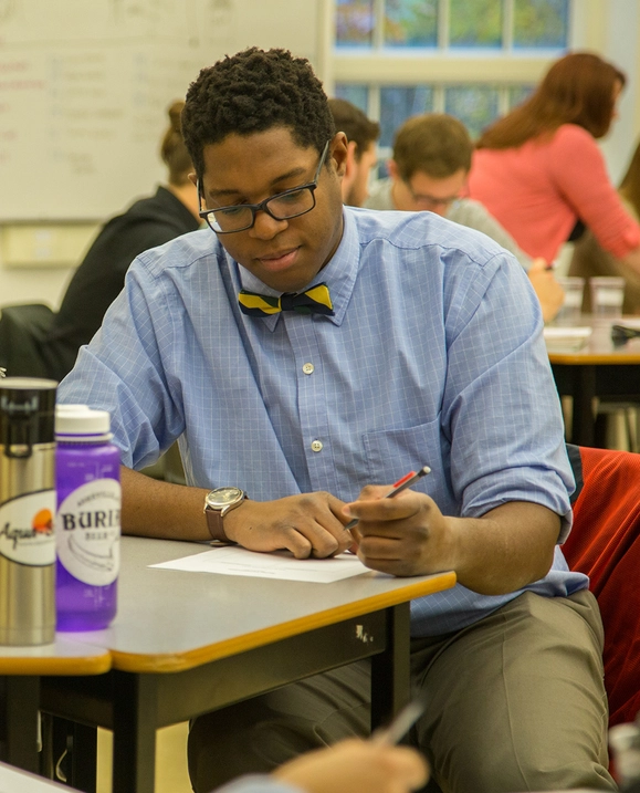A male student wearing a dress shirt and bow tie sits at a table doing work