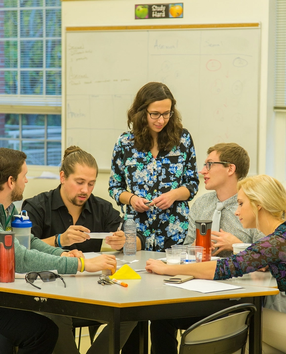 A female student stands beside a table of her peers, instructing them on a class lesson.