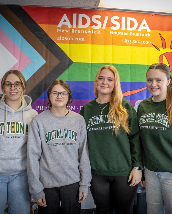 A group of students wearing St. Thomas Social Work sweaters stand in front of an AIDS New Brunswick pride flag