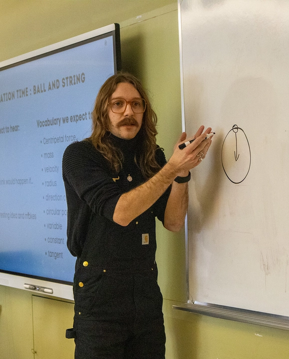 A male teacher candidate stands at a whiteboard holding a dry-erase marker as he instructs the class