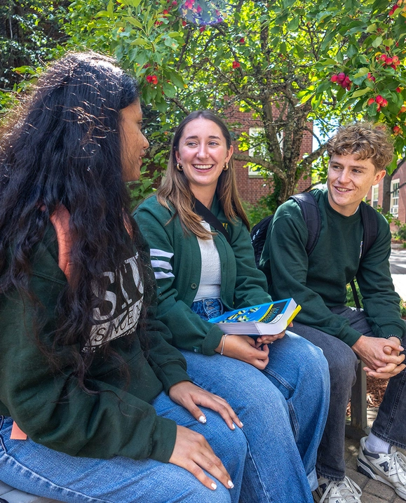 Three students—two female and one male—sit on a bench outside on campus, chatting with one another.