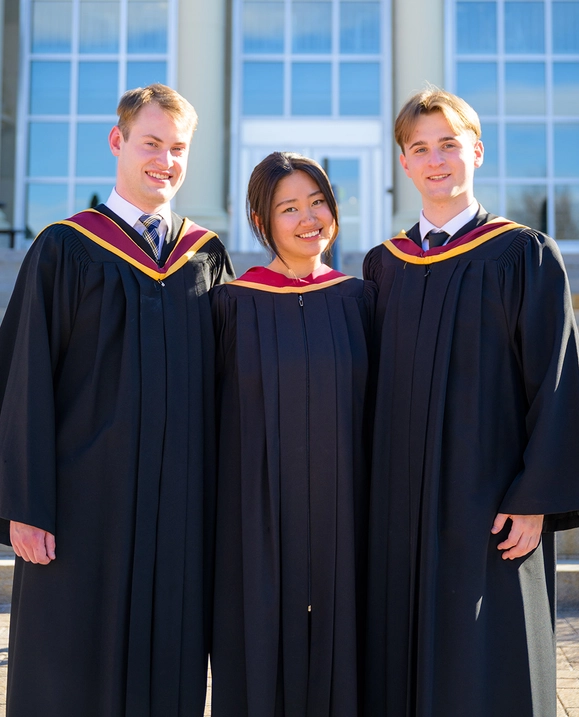 Three STU graduates stand together in front of George Martin Hall wearing their grad gowns
