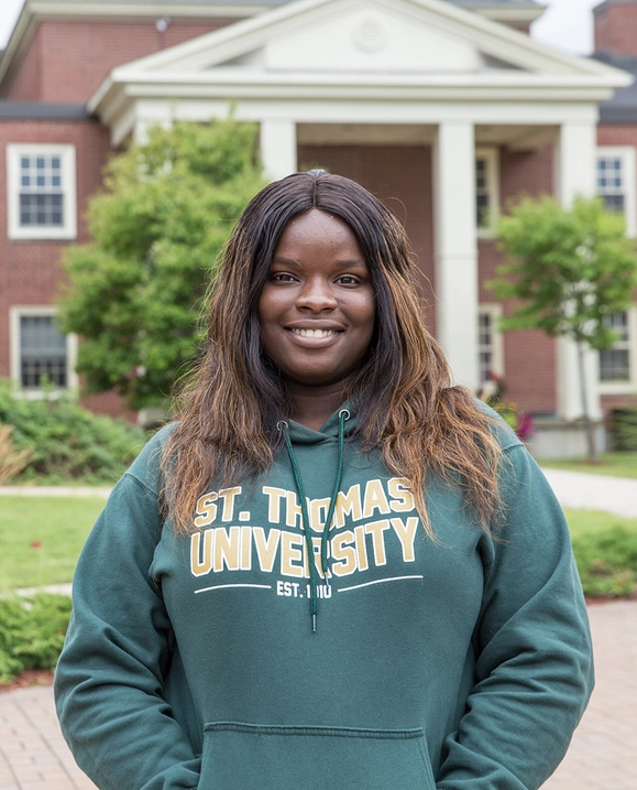 A female student stands on campus wearing a green St. Thomas University sweater