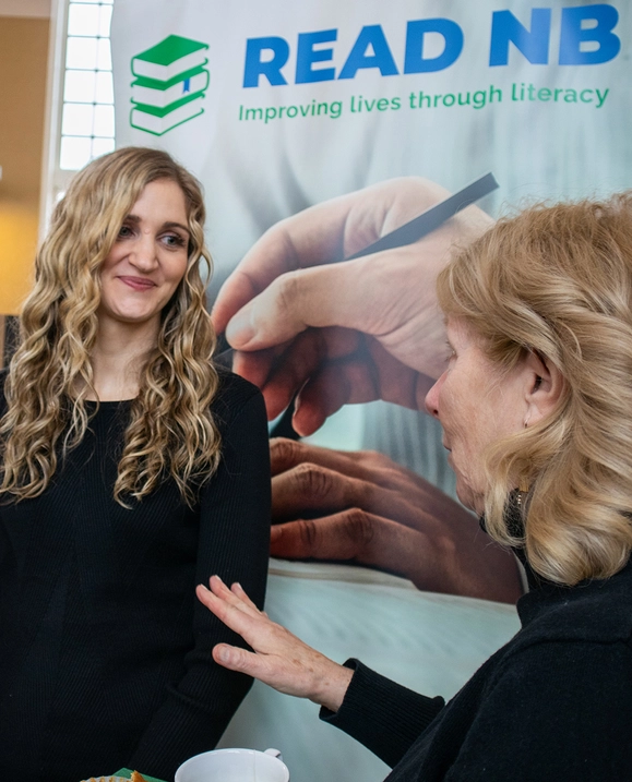 A female student stands in front of a Literacy New Brunswick banner, talking with a woman