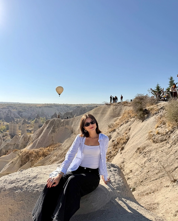A female student sits on a sandy landscape with a hot air balloon flying in the background