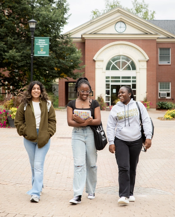 Three female students walk through campus holding books with Duffie Hall in the background
