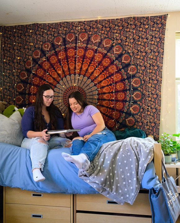 Two students sit on a bed in residence, looking over a notebook
