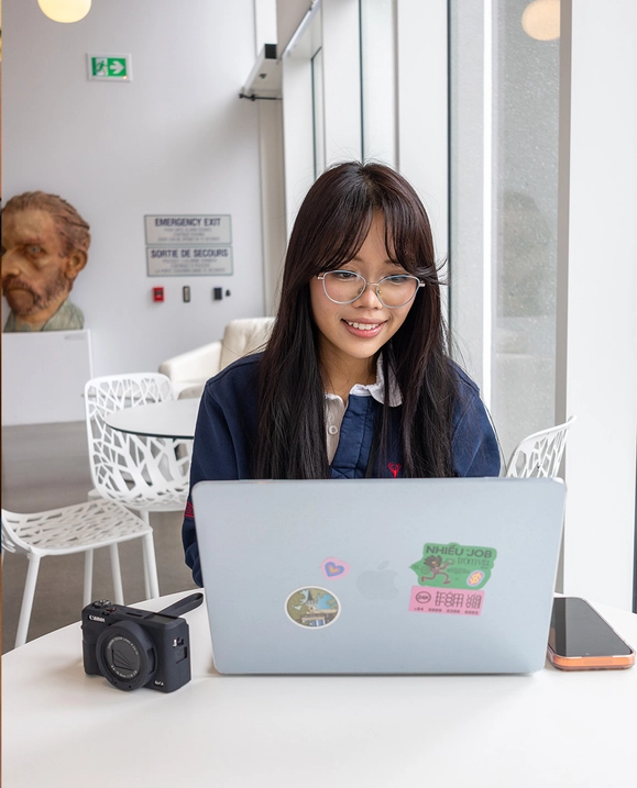 A female student sits at a white table with her laptop, camera, and cell phone.