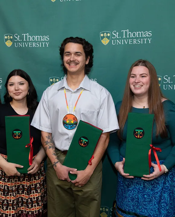 Two female students and one male student stand in front of a green St. Thomas backdrop holding their Eagle Feather folders