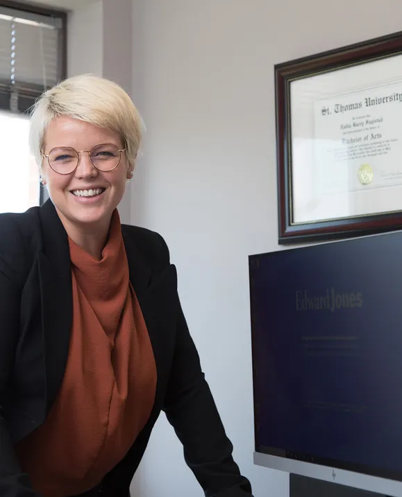 A female alumnus stands behind her desk in her office with her STU degree hanging behind her