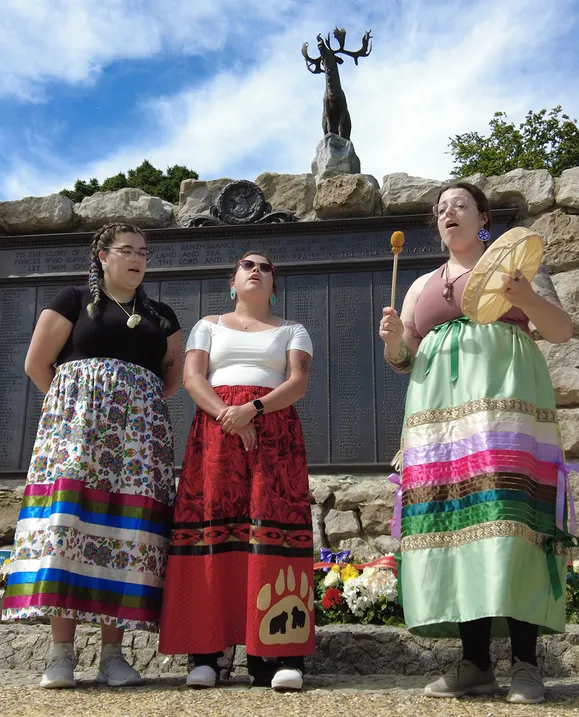 Three indigenous women wearing ribbon skirts sing and drum in front of a memorial site
