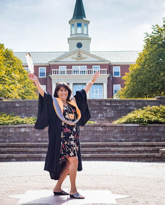 A female graduate stands in lower campus in her grad gown, proudly raising her eagle feather in the air