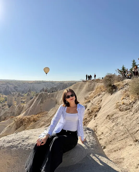 A female student sits on a sandy landscape with a hot air balloon flying in the background