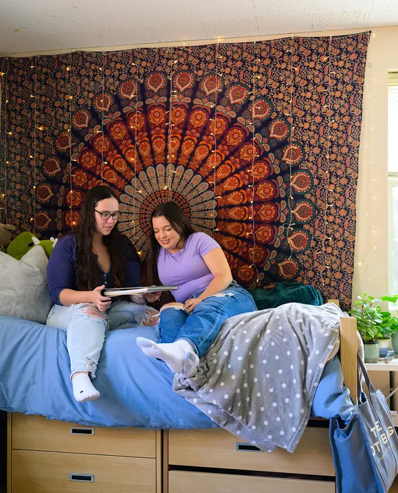 Two students sit on a bed in residence, looking over a notebook