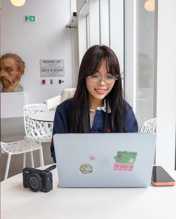 A female student sits at a white table with her laptop, camera, and cell phone.