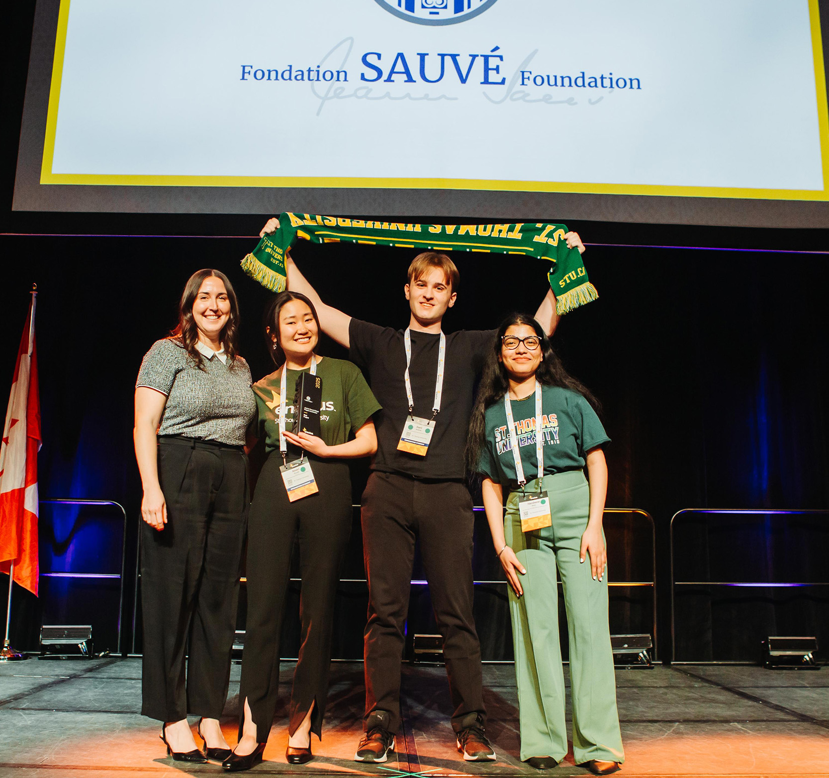 A female student holding an award, a male student holding up a green and gold ߲ݴý scarf, and another female student stand on a stage