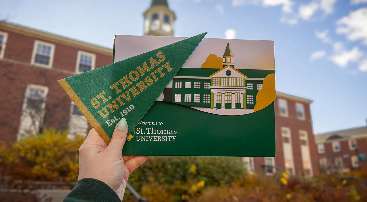 A hand holding out a STU pennant and an Acceptance Folder in front of George Martin Hall