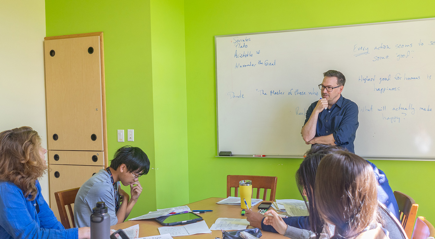 Dr. Andrew Moore stands in front of a white board with writing on it as a student at a table talks with him during a class