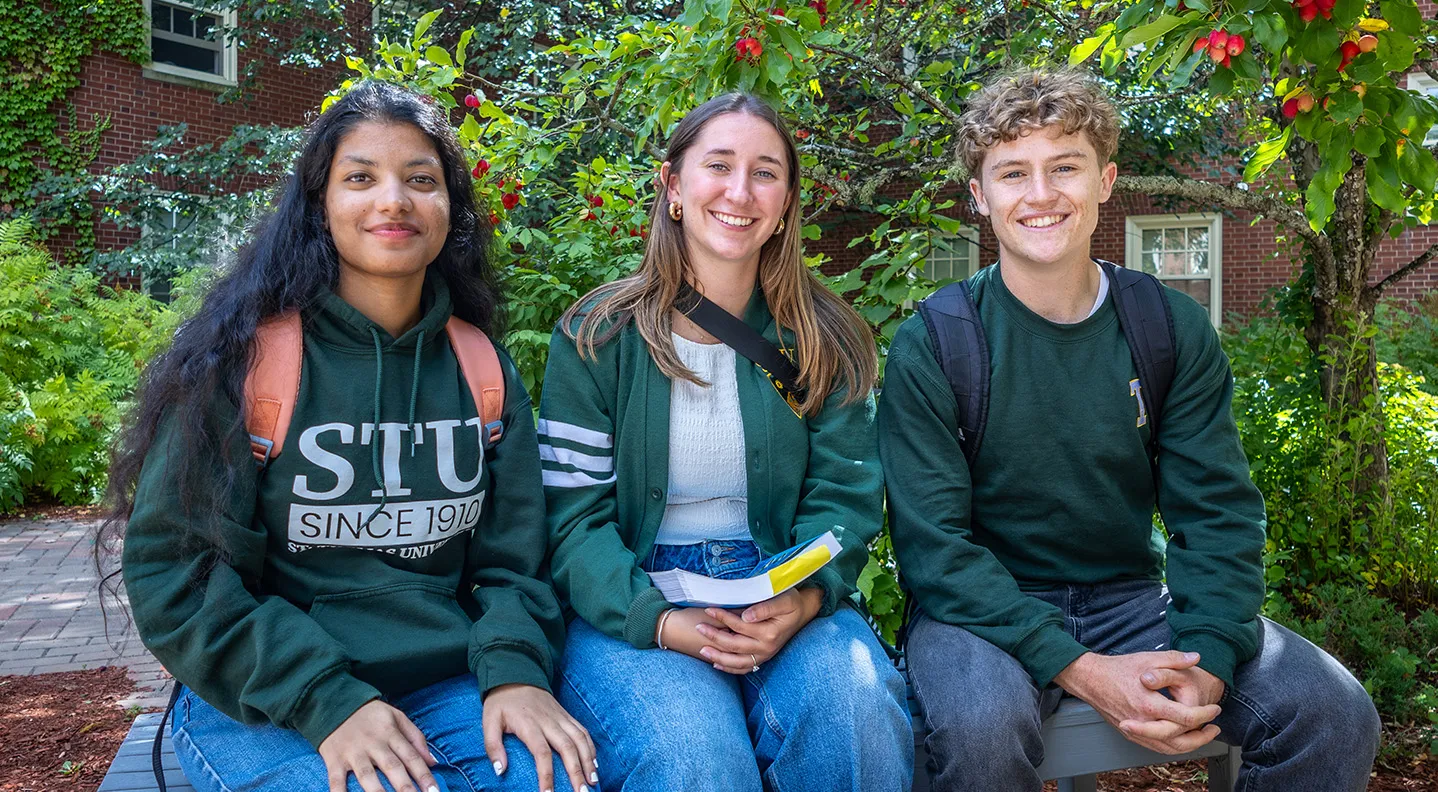 Two female students and one male student sitting on a bench outside on campus
