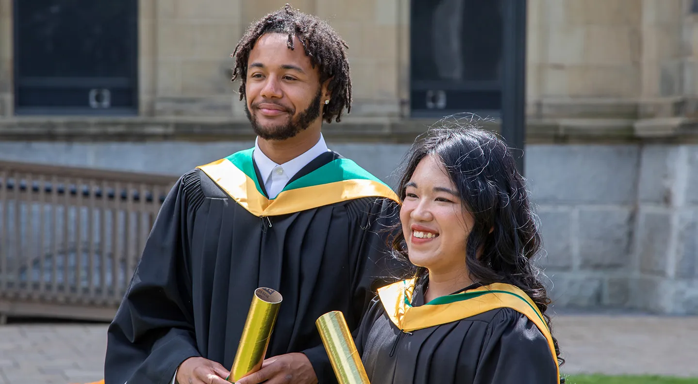 Two Master of Social Work grads stand together in their gowns holding their degrees