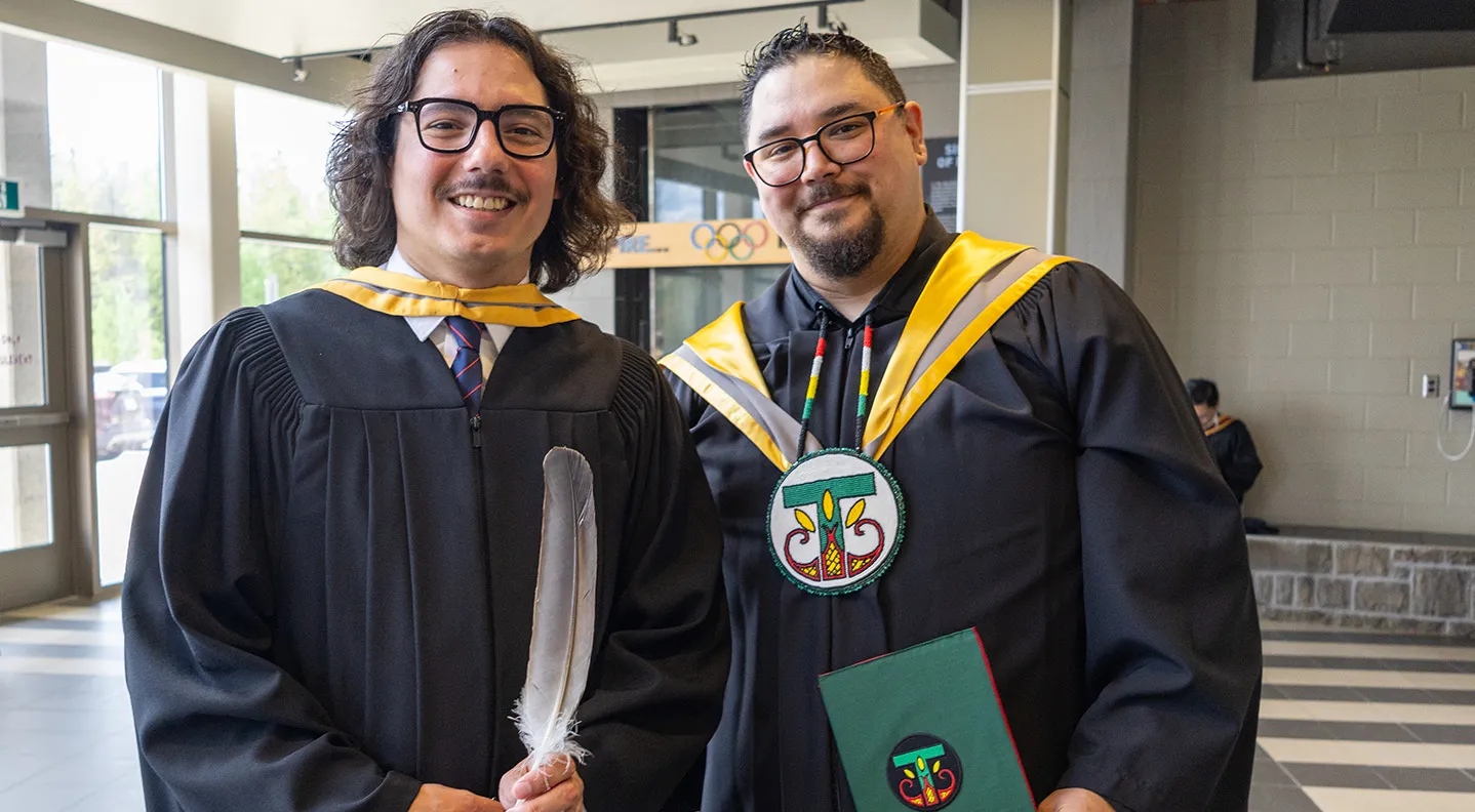 Two male Indigenous graduates stand together holding the Eagle Feathers they received prior to Spring Convocation