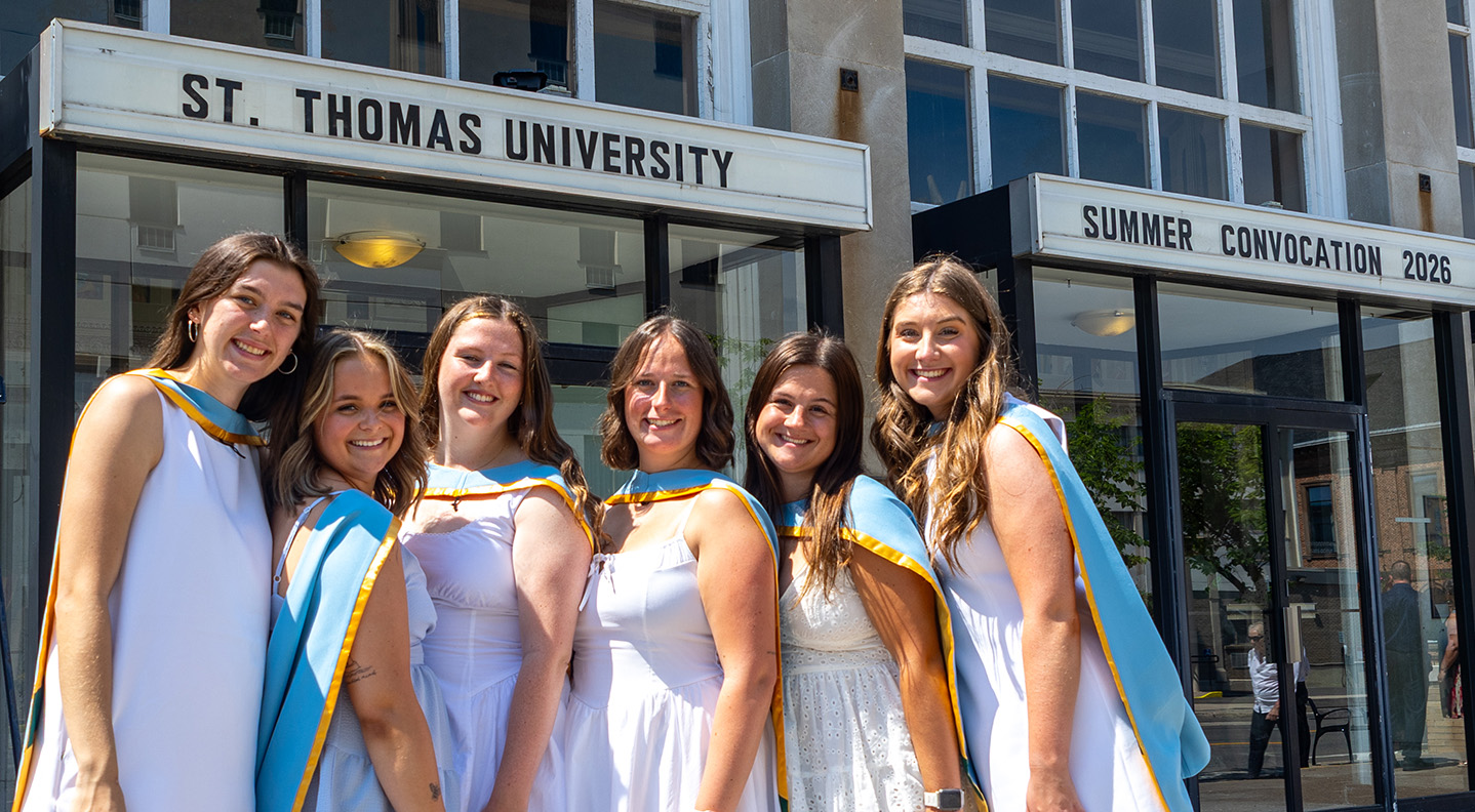 A group of female graduates stand with their Bachelor of Education hoods on in front of the Fredericton Playhouse where the marquee reads 