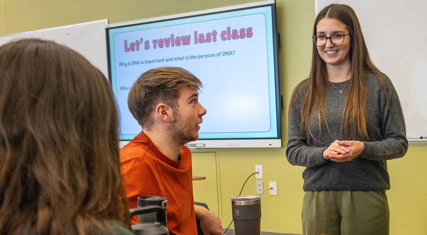 A female teacher candidate stands at the front of the class discussing her lesson with her peers